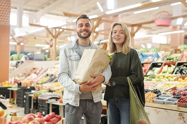 Retrato de um jovem casal positivo em pé no mercado dos fazendeiros e comprando alimentos orgânicos juntos