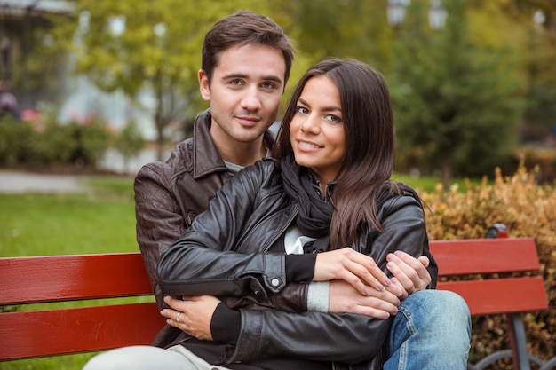 Retrato de um jovem casal sorridente, sentado no banco ao ar livre e olhando para a frente