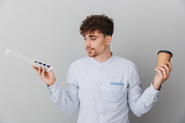 Retrato de um jovem com a barba por fazer, vestido com uma camisa e expressando hesitação, segurando um café para viagem e um jornal nas mãos, isolado sobre a parede cinza