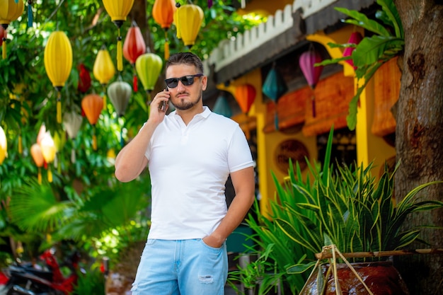 Retrato de um jovem com óculos, caminhando ao ar livre com um telefone celular. Um homem com um telefone. Uma pessoa feliz está falando ao telefone.