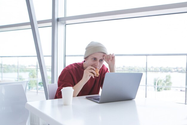 Retrato de um jovem comendo um sanduíche em um café e olhando para uma tela de laptop