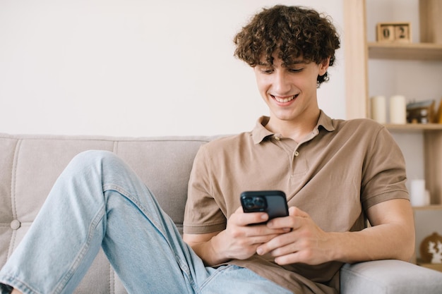 Retrato de um jovem de cabelo encaracolado feliz usando smartphone enquanto está sentado no sofá na sala de estar e descansando