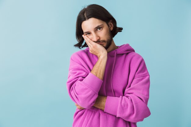 Foto retrato de um jovem deprimido, barbudo, moreno, vestindo um moletom, isolado na parede azul