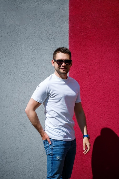 Retrato de um jovem elegante, um homem vestido com uma camiseta branca em branco de pé sobre um fundo de parede cinza e vermelho. estilo urbano de roupas, imagem na moda moderna. moda masculina