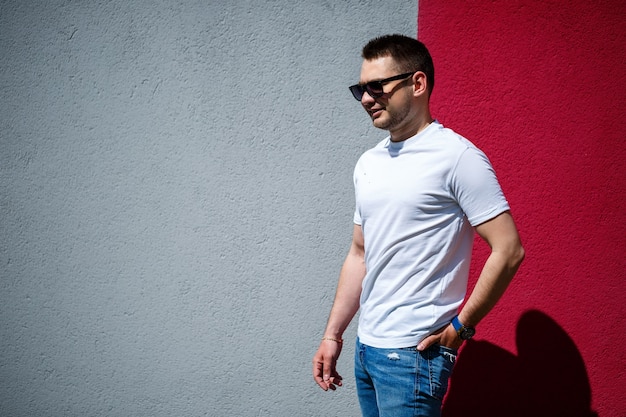 Retrato de um jovem elegante, um homem vestido com uma camiseta branca em branco de pé sobre um fundo de parede cinza e vermelho. Estilo urbano de roupas, imagem na moda moderna. Moda masculina