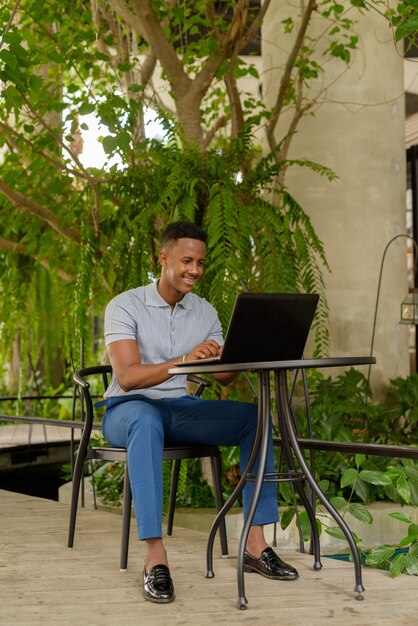 Retrato de um jovem empresário africano feliz vestindo roupas casuais e sentado na cafeteria enquanto usa o computador laptop e o distanciamento social