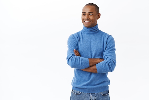 Retrato de um jovem empresário afro-americano de sucesso sonhador em gola alta azul cruzar as mãos sobre o peito desviar o olhar do espaço de cópia do lado esquerdo com fundo branco de expressão satisfeito satisfeito