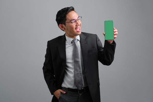 Retrato de um jovem empresário alegre e bonito em terno formal mostrando smartphone com tela vazia isolada em fundo cinza