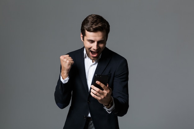 Retrato de um jovem empresário animado vestido com um terno isolado sobre uma parede cinza, segurando um telefone celular, comemorando