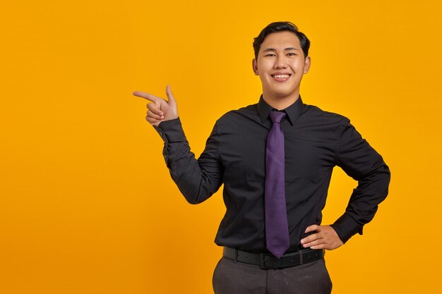 Retrato de um jovem empresário asiático sorridente apontando o dedo para o espaço da cópia em fundo amarelo