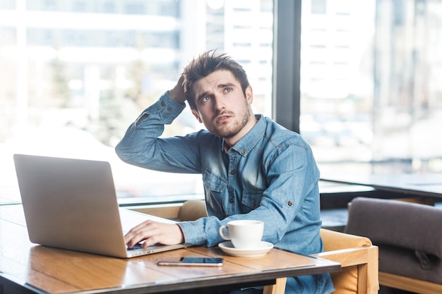 Retrato de um jovem empresário barbudo pensativo bonito em camisa jeans azul está sentado no café e digitando a mensagem no laptop, tendo uma nova ideia e planejando a própria estratégia, segurando uma mão na cabeça.