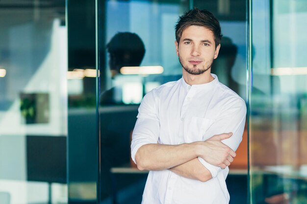 Retrato de um jovem empresário bonito freelancer de pé no escritório em roupas brancas braços cruzados olhando para a câmera sorrindo