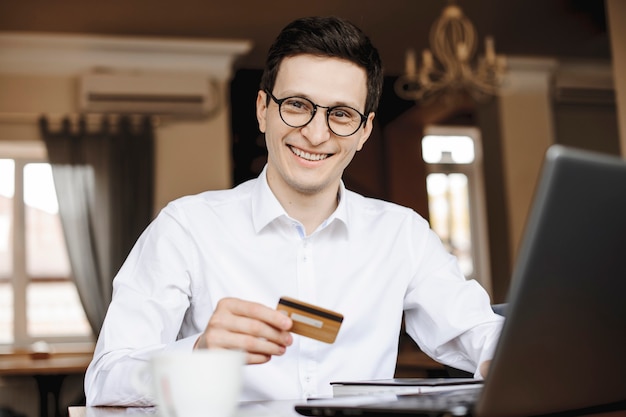 Retrato de um jovem empresário bonito, olhando para a câmera rindo, segurando um cartão de crédito ouro, sentado em uma mesa trabalhando em seu laptop.