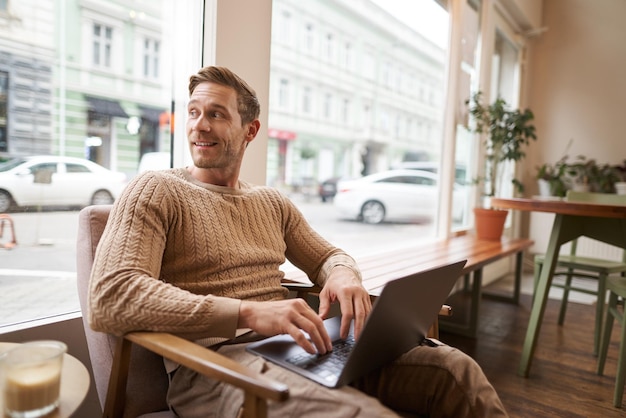 Retrato de um jovem empresário bonito, um homem com um laptop sentado em uma cafeteria em uma cadeira e trabalhando