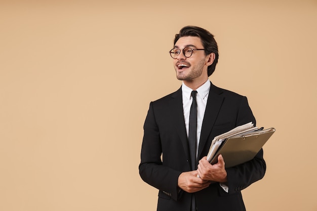 Retrato de um jovem empresário confiante e sorridente, vestindo um terno de pé isolado sobre uma parede bege, segurando uma pilha de documentos