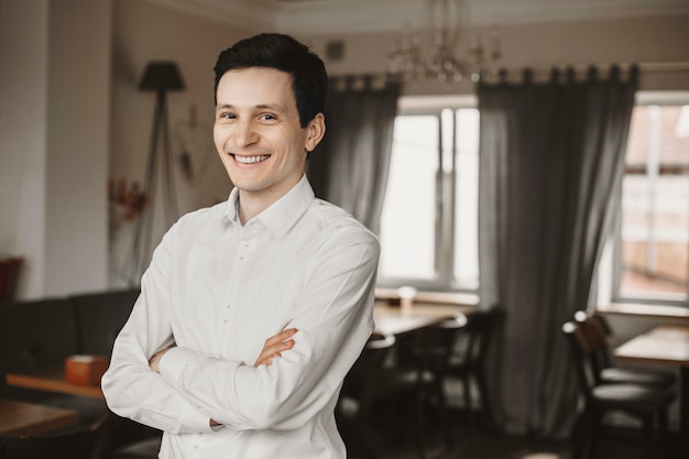 Retrato de um jovem empresário confiante sentado em seu restaurante com as mãos cruzadas e olhando para a câmera sorrindo.