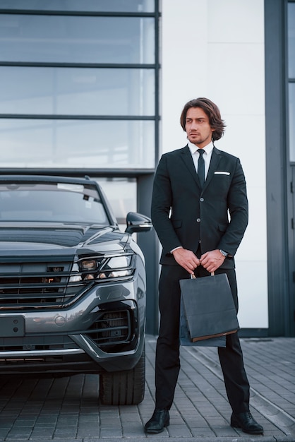 Retrato de um jovem empresário de terno preto e gravata ao ar livre perto de um carro moderno e com sacolas de compras.