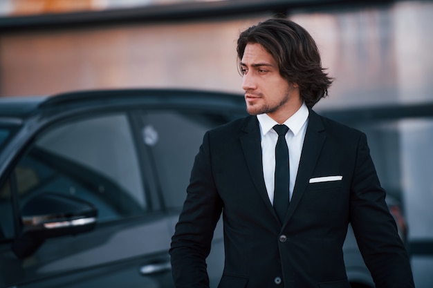 Retrato de um jovem empresário de terno preto e gravata ao ar livre perto de um carro moderno.