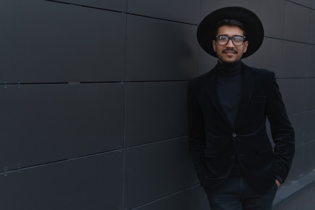 Retrato de um jovem empresário descolado de terno preto e chapéu perto do prédio da parede, copyspace