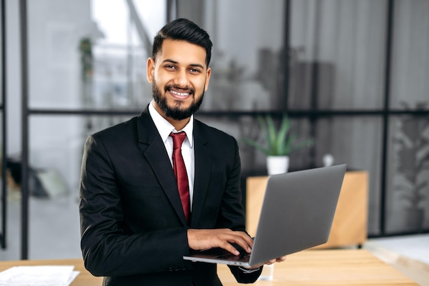 Retrato de um jovem empresário indiano ou árabe elegante e confiante bem-sucedido de terno segurando um laptop aberto na mão perto da área de trabalho em seu escritório moderno, olhando para a câmera sorrindo