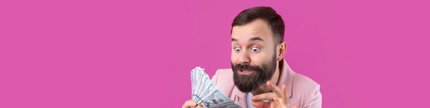 Retrato de um jovem empresário satisfeito com uma barba vestida com uma jaqueta rosa mostrando notas de dólar contra um fundo de estúdio vermelho Gosto de cheiro de dinheiro