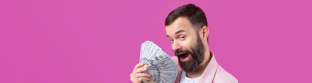 Retrato de um jovem empresário satisfeito com uma barba vestida com uma jaqueta rosa mostrando notas de dólar contra um fundo de estúdio vermelho Gosto de cheiro de dinheiro