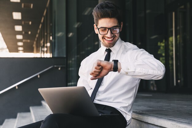 Retrato de um jovem empresário vestido com um terno formal sentado do lado de fora de um prédio de vidro com um laptop e olhando para seu relógio de pulso