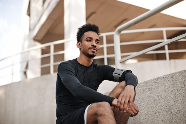 Retrato de um jovem encaracolado, barbudo, de pele escura, com camiseta preta de mangas compridas e shorts, sentado na escada do lado de fora