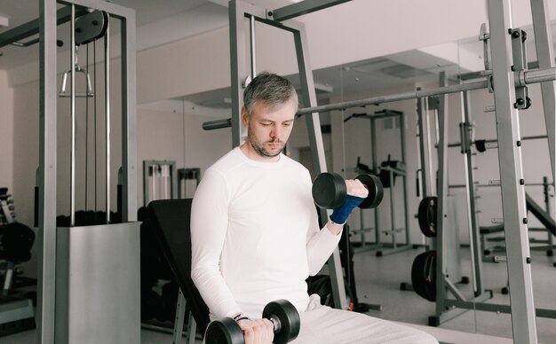 Retrato de um jovem fazendo exercícios físicos, treinamento de bíceps com halteres no ginásio, uma cópia do espaço. Em uma camiseta branca.