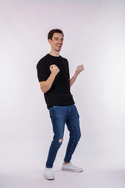 Retrato de um jovem feliz e alegre em pé fazendo gesto de vencedor, cerrando os punhos, mantendo-se isolado no estúdio de fundo de parede de cor branca