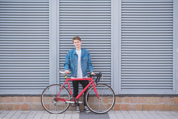 Retrato de um jovem feliz e elegante em uma jaqueta jeans e uma bicicleta vermelha em uma parede cinza