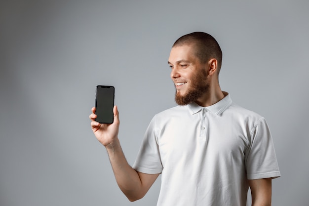 Retrato de um jovem feliz em uma camiseta branca, mostrando uma tela de telefone em cinza.