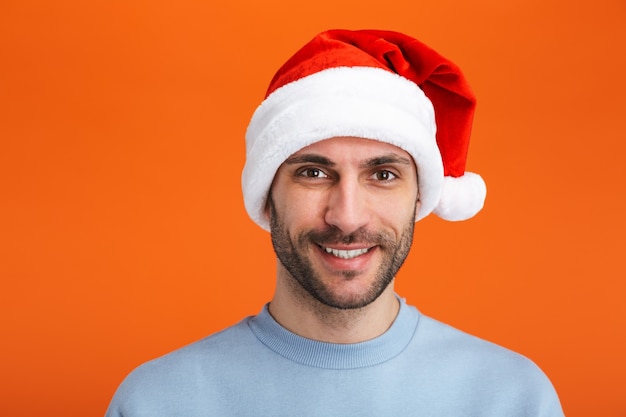 Retrato de um jovem feliz otimista positivo no chapéu de Papai Noel de Natal posando isolado sobre uma parede laranja.