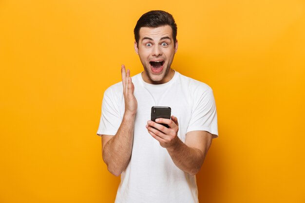 Retrato de um jovem feliz vestindo uma camiseta em pé, isolado na parede amarela, usando telefone celular, comemorando o sucesso