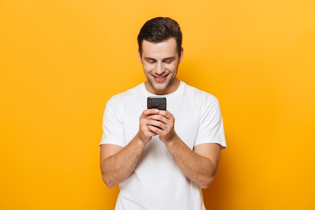 Retrato de um jovem feliz vestindo uma camiseta em pé, isolado na parede amarela, usando telefone celular