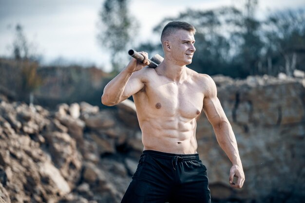 Retrato de um jovem fisiculturista com tubo de metal em um ombro nu torso Quarry background