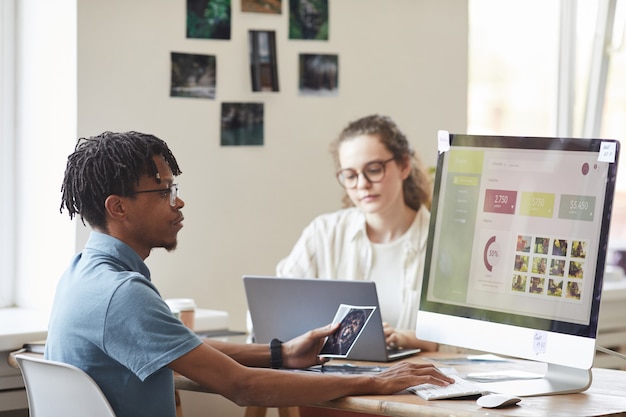 Retrato de um jovem fotógrafo afro-americano gerenciando o mercado de ações de fotos enquanto usa o computador na mesa do escritório com o site na tela, copie o espaço