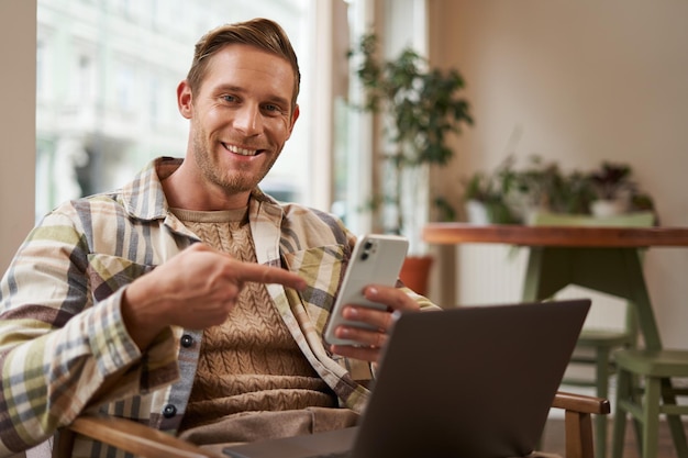 Retrato de um jovem freelancer trabalhando em um café sentado em uma cadeira com um laptop apontando o dedo para