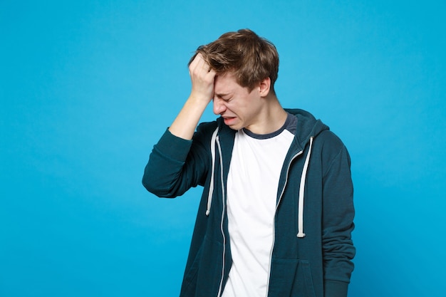 Retrato de um jovem frustrado a chorar em roupas casuais, com a cabeça baixa, colocando a mão na cabeça isolada na parede azul. conceito de estilo de vida de emoções sinceras de pessoas.