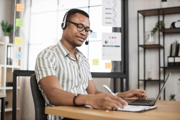 Foto retrato de um jovem gerente de escritório africano sorridente em camisa listrada e relatório de redação de fone de ouvido