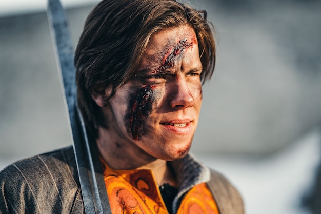 Retrato de um jovem guerreiro medieval em armadura com espada no ombro. No processo de batalha, com feridas no rosto