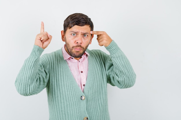 Retrato de um jovem homem mostrando um gesto de eureka, apontando para as têmporas com uma camisa, um casaco de lã e uma vista frontal inteligente