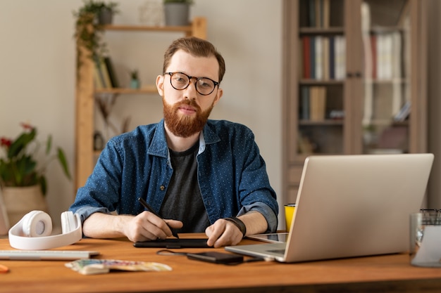 Retrato de um jovem ilustrador masculino bonito em óculos, sentado à mesa com um bloco de desenho e um laptop no escritório em casa