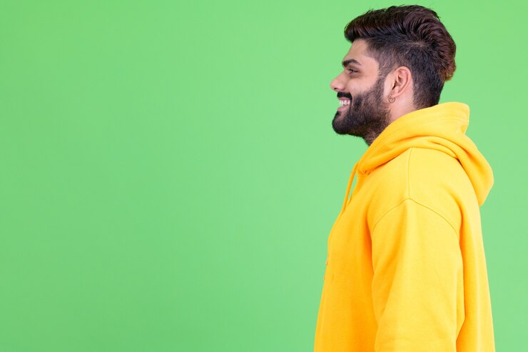 Foto retrato de um jovem indiano barbudo com excesso de peso usando um capuz contra o chroma key com uma parede verde