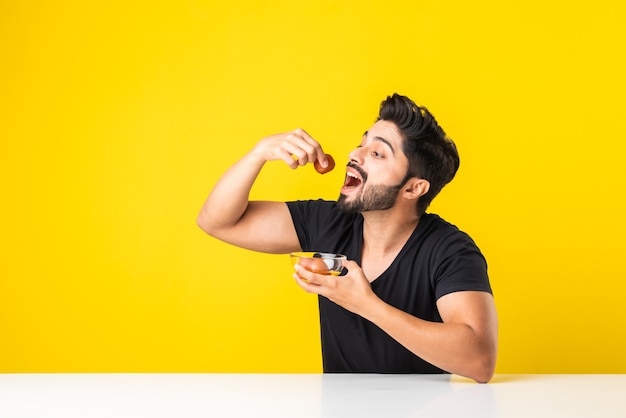 Retrato de um jovem indiano bonito comendo o doce Gulab Jamun contra um fundo amarelo