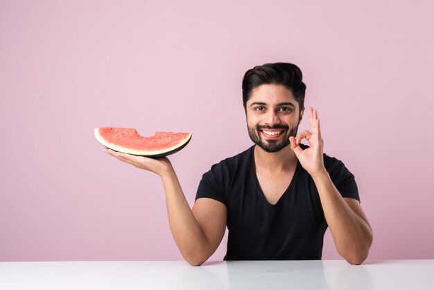 Retrato de um jovem indiano bonito sorrindo e comendo melancia fresca ou tarbooj enquanto está sentado à mesa ou isolado no chão de madeira