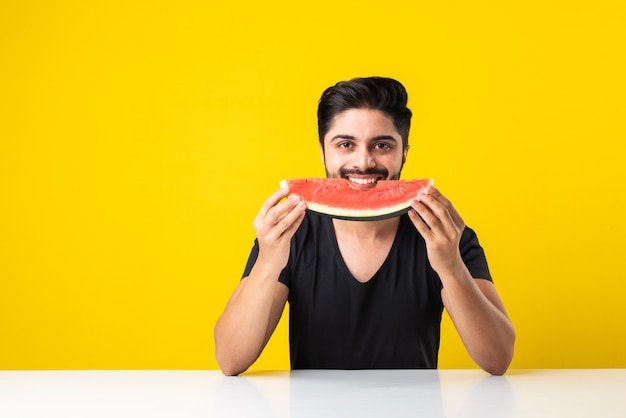 Retrato de um jovem indiano bonito sorrindo e comendo melancia fresca ou tarbooj enquanto está sentado à mesa ou isolado no chão de madeira