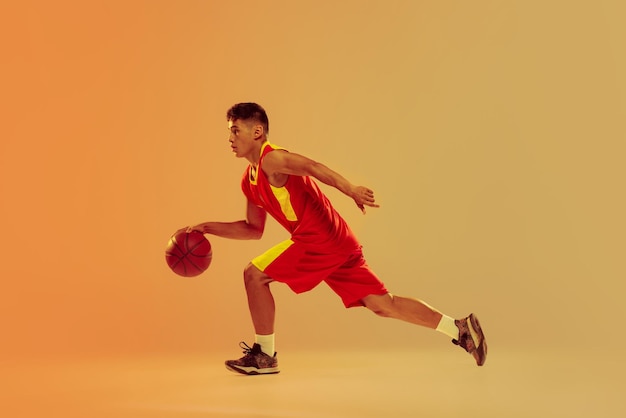 Retrato de um jovem jogador de basquete treinando driblando isolado sobre o fundo laranja do estúdio em