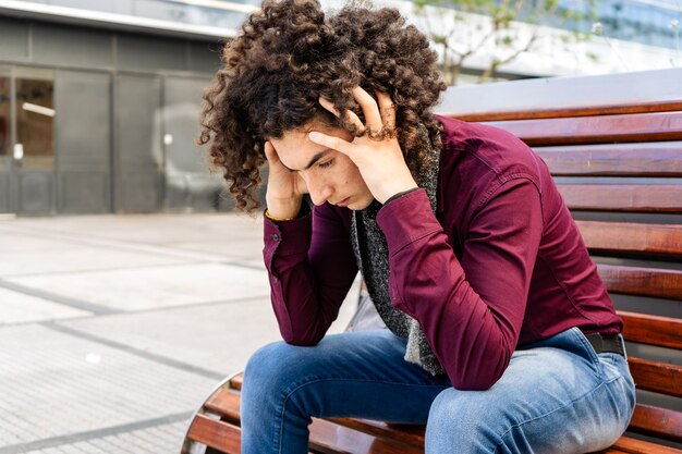 Retrato de um jovem latino com cabelos cacheados, sentado em um banco na rua, segurando sua cabeça, mostrando frustração ou preocupação.