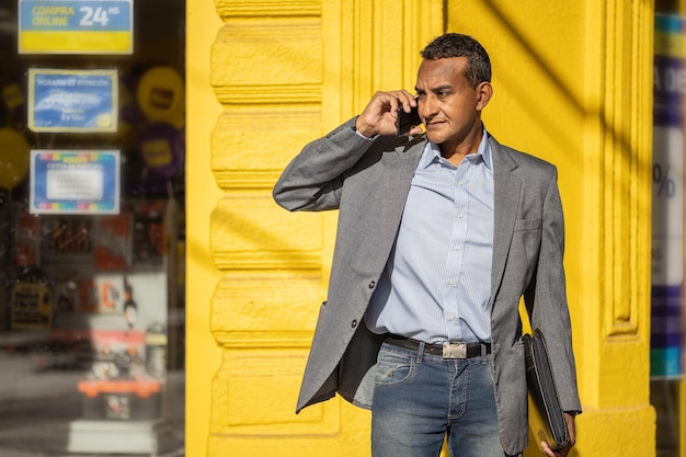 Retrato de um jovem latino falando no celular sobre uma parede amarela